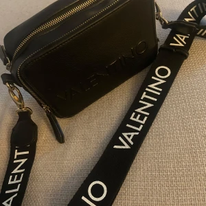 Svart axelväska från Valentino - Snygg svart axelväska från Valentino med justerbar axelrem och logotyp i relief. Väskan har två dragkedjor.