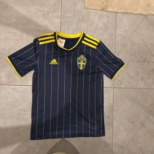 Blå och gul Sverige fotbollströja från Adidas - Snygg blå fotbollströja från Adidas med gula ränder och Sveriges emblem på bröstet. Tröjan har korta ärmar och en rund gul krage. Perfekt för att heja på landslaget! 🇸🇪