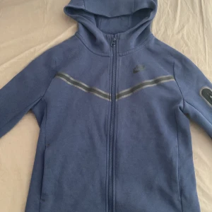 Nike tech - Säljer en blå huvtröja från Nike med dragkedja och huva. Tröjan har ett diskret märke på bröstet och en reflekterande rand över bröstet. Perfekt för en sportig look!