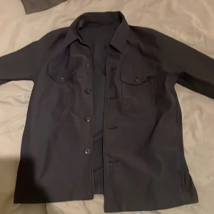 Svart overshirt med knappar - Säljer en stilren svart overshirt med knappar framtill. Den har långa ärmar och två bröstfickor med knappar. Perfekt för lager-på-lager-stil. Passar till många olika outfits! Bara använd 1-2 gånger så inprincipt helt ny. Storlek  S men passar även M