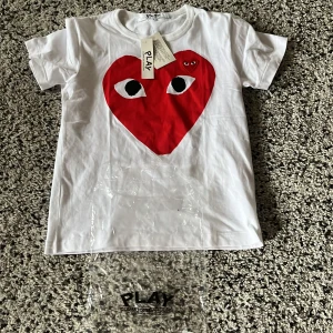 Vit t-shirt med rött hjärta från Comme des Garçons - Säljer en vit t-shirt från Comme des Garçons med ett stort rött hjärta med ögon på framsidan. T-shirten är kortärmad och har en klassisk passform. Perfekt för en stilren och lekfull look. Passar någon som är 170-178. Den är helt nya med tags. Säljer pga den var för liten
