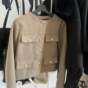 Beige kavaj från Zara - Snygg beige kavaj från Zara med guldknappar och fyra fickor framtill. Den har en klassisk design med rund hals och långa ärmar. Perfekt för en stilren look.