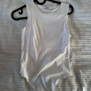 Vit ärmlös bodysuit från NA-KD - Säljer en stilren vit ärmlös bodysuit från NA-KD i storlek XS. Perfekt för lager-på-lager eller att bära som den är. Den har en enkel knäppning i grenen för smidig användning. Passar till många olika outfits och tillfällen.