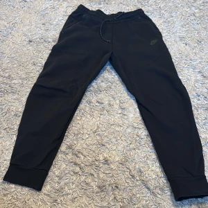 Svarta mjukisbyxor från Nike - Snygga svarta mjukisbyxor från Nike med elastisk midja och snörning. Byxorna har en dragkedja på ena sidan och är perfekta för en avslappnad stil. De har en normal passform och är tillverkade i ett mjukt material för extra komfort.