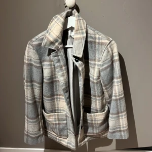 Grå rutig kappa - Snygg grå rutig kappa med klassisk krage och knappar framtill. Jackan har två stora fickor och en stilren design som passar perfekt för kyligare dagar. Perfekt för dig som vill ha en stilren look med en touch av klassisk elegans.