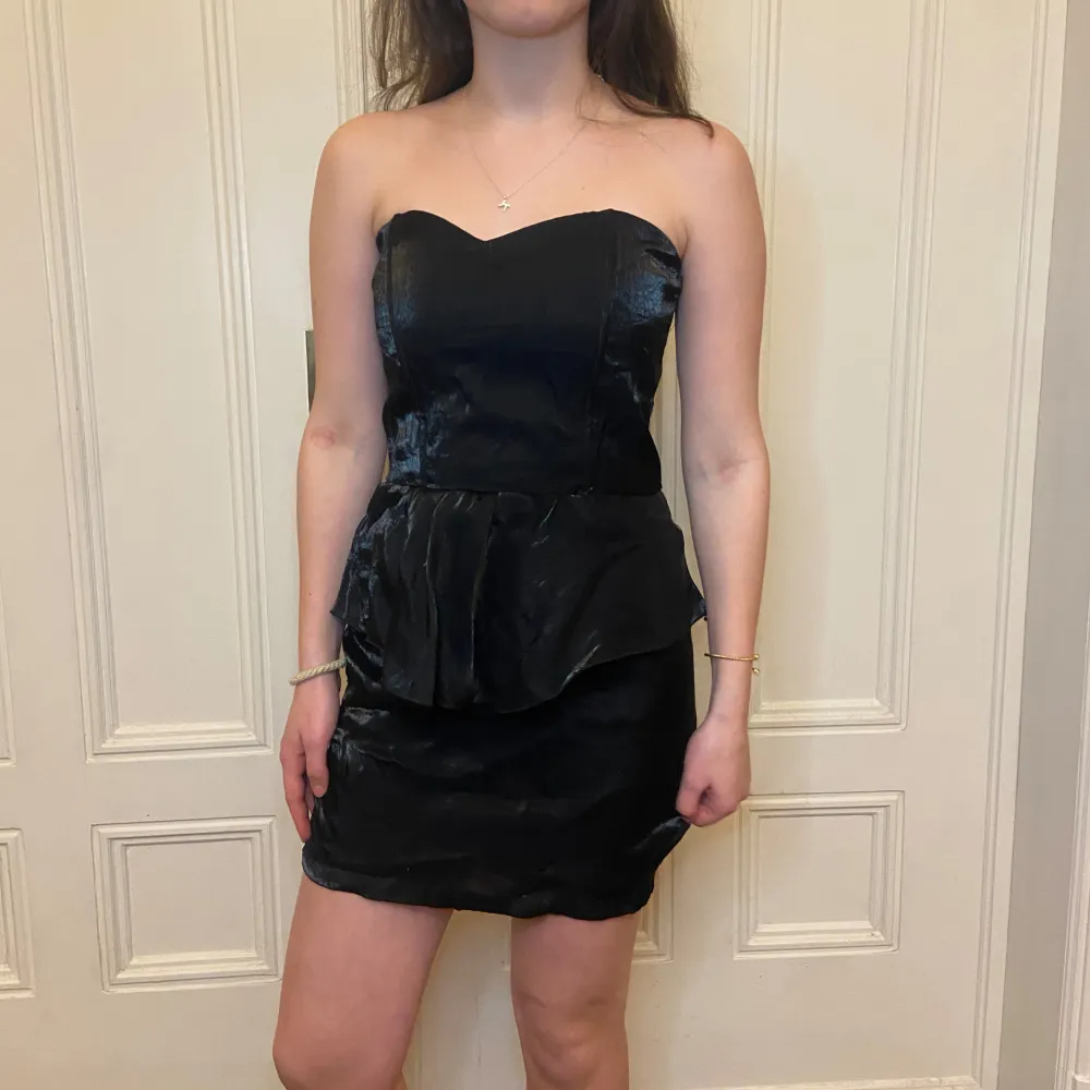 Snygg svart axelbandslös klänning med en peplumdetalj i midjan. Klänningen har en dragkedja i ryggen och är perfekt för en kväll ute. Den korta längden ger en chic look. Köpt second hand!!. Mekot.