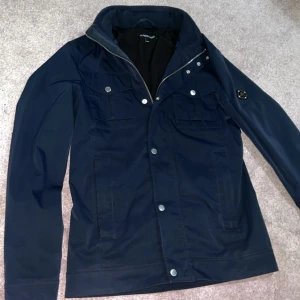 Mörkblå jacka från J.Lindeberg - Snygg mörkblå jacka från J.Lindeberg med dragkedja och knappar. Jackan har en stilren design med två bröstfickor och en hög krage. Perfekt för en stilren look.