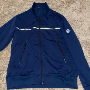 Moncler kofta med dragkedja - Snygg mörkblå kofta från Moncler med dragkedja framtill och två fickor med dragkedjor. Koftan har en hög krage och ett reflekterande band över bröstet och ryggen. Perfekt för en stilren look. Pris kan diskuteras. 😃