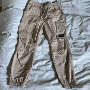 Beige cargobyxor med elastisk midja - Snygga beige cargobyxor med elastisk midja och muddar vid bensluten. Byxorna har flera praktiska fickor, inklusive stora sidofickor med lock. Perfekta för en avslappnad stil.
