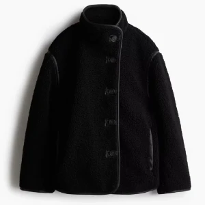 Svart teddyjacka med knappar - Snygg svart teddyjacka med hög krage och knappar framtill. Jackan har en mysig och varm känsla, perfekt för kyliga dagar. Den har långa ärmar och en stilren design som passar till många olika outfits.