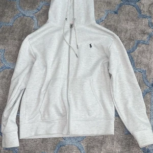 Grå hoodie från Polo Ralph Lauren - Säljer en stilren grå hoodie från Polo Ralph Lauren med dragkedja och klassisk logga på bröstet. Tröjan har en bekväm passform med långa ärmar och en praktisk huva med snörning. Perfekt för en avslappnad look.