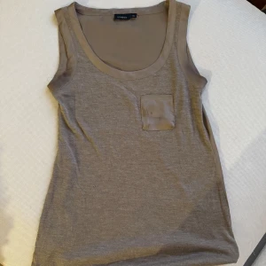 Beige linnetopp från Lindex - Säljer en stilren beige linnetopp från Lindex. Toppen har en liten bröstficka och är ärmlös, perfekt för varma dagar. Den är gjord i ett mjukt och bekvämt material.