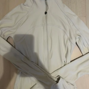Beige jacka från Lululemon - !INTRESEKOLL!Säljer en beige jacka från Lululemon med dragkedja framtill. Jackan har en slim passform och är tillverkad i ett syntetmaterial med ribbade detaljer. Perfekt för en stilren look. *tryck ej på köp nu*