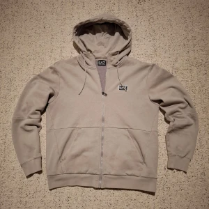Beige hoodie från Emporio Armani - Säljer en stilren beige hoodie från Emporio Armani med dragkedja och huva. Tröjan har en diskret logga på bröstet och är tillverkad i ett mjukt material. Perfekt för en avslappnad look.