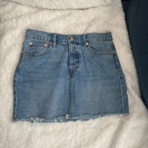 Jeanskjol från Lager 157 - Snygg jeanskjol från 157 i klassisk blå denim. Kjolen har en kort längd med rå kant nertill och femficksdesign. Den stängs med knappar framtill och har bälteshällor för extra stil. Perfekt för en avslappnad look!