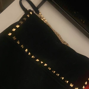 Svart axelväska med guldkedja - Snygg svart axelväska med guldkedja och nitar. Väskan har en dragkedja och är perfekt för att ge en edgy touch till din outfit. Den är både stilren och praktisk med sina detaljer. Stockholm stil väska. 