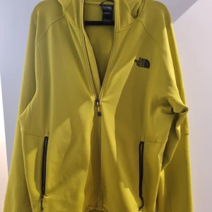 Fleecejacka från The North Face - Fleecejacka från The North Face med dragkedja och huva. Jackan har långa ärmar och två fickor med dragkedjor framtill. Tunnare modell.
