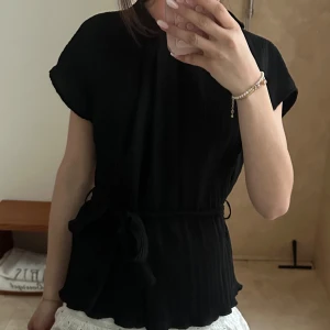 Svart blus med knytband - Elegant svart blus med korta ärmar och ett knytband i midjan. Blusen har en subtil ribbad textur och en feminin siluett som passar perfekt för både vardag och speciella tillfällen. Från zara och super skick!