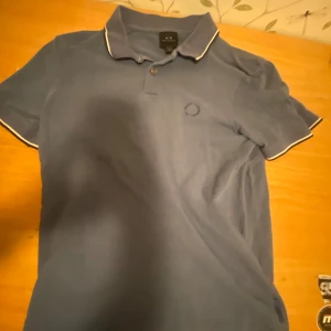 Blå pikétröja från Armani Exchange - Snygg blå pikétröja från Armani Exchange med vit kant på krage och ärmar.  Lite sliten på loggan men annars bra skick (pris kan diskuteras vid snabb affär 