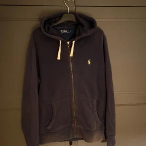 Mörkblå hoodie från Ralph Lauren med zip - Säljer en snygg mörkblå hoodie från Ralph Lauren med dragkedja och huva. Tröjan har en liten broderad logga på bröstet och praktiska fickor framtill. Perfekt för en avslappnad stil.