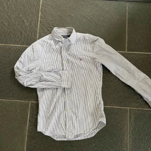 Randig skjorta från Ralph Lauren - Snygg vit och blå randig skjorta från Ralph Lauren med klassisk krage och knappar framtill. Skjortan har långa ärmar och en liten broderad logga på bröstet. Perfekt för en stilren look!
