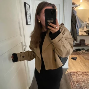Beige croppad trenchcoat - Säljer en beige croppad trenchcoat med knappar och långa ärmar. Jackan har en klassisk krage och är perfekt för en stilren look. Passar bra till både jeans och klänning.