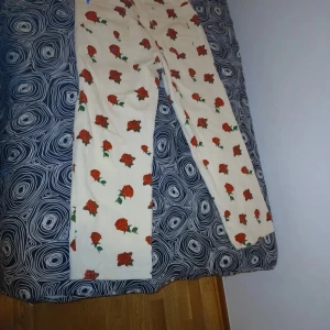 Vita byxor med rosor - Unika vita byxor med ett vackert ros-mönster i rött och grönt. Byxorna har en rak passform och är perfekta för att sticka ut med sin blommiga design. De är både stilrena och lekfulla.