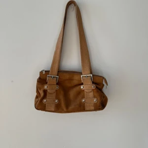 Brun axelväska från Andrea - Säljer en snygg brun axelväska från Andrea i mocka med coola metalldetaljer och dragkedja. Väskan har en rymlig insida med flera fack för enkel organisering. Perfekt för att bära med sig det viktigaste.