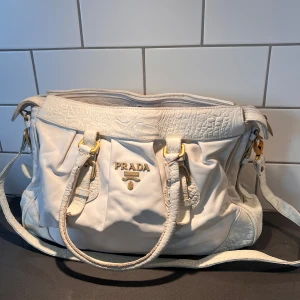 Vit axelväska  - Säljer en stilren vit axelväska  i skinn. Väskan har eleganta gulddetaljer och flera praktiska fack inuti. Perfekt för att bära med sig allt man behöver. Den har både handtag och en justerbar axelrem för flexibilitet.Väskan får utrymme liten arbetsstol, motto fixan får plats IPad och A4 block . 