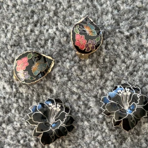 Svarta örhängen med blommönster - Säljer ett par unika svarta örhängen med vackra blommönster i färgerna rosa, blå och guld. Örhängena har en elegant design med detaljerade blommor och fjärilar, perfekt för att ge en färgklick till din outfit. 175 kr för både par