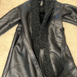 Svart kappa från Boohoo - Snygg svart kappa från Boohoo med fuskpälsdetaljer. Den har en lång och elegant design med bälte i midjan för en smickrande passform. Perfekt för kyligare dagar när du vill hålla stilen.