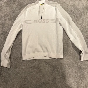 Vit half zip från Boss unik!! - Säljer en stilren vit tröja från Boss med dragkedja vid halsen. Tröjan har långa ärmar och ett subtilt mönster med Boss-loggan över bröstet. Perfekt för en avslappnad men snygg look.
