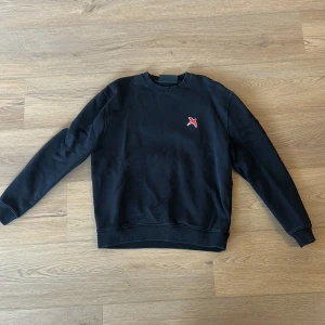 Svart sweatshirt med röd logga - Säljer en stilren svart sweatshirt från Axel Arigato med en liten röd fågel på bröstet. Tröjan har långa ärmar och ribbade muddar vid ärmslut och nederkant. Perfekt för en avslappnad look.