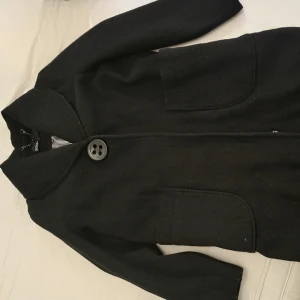 Svart kappa - Snygg svart kappa med stora knappar framtill. Den har en klassisk krage och två stora fickor. Perfekt för kyligare dagar och ger en stilren look.
