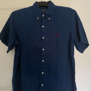 Blå kortärmad skjorta från Polo Ralph Lauren - Snygg blå kortärmad skjorta från Polo Ralph Lauren med klassisk krage och knappar framtill. Den har en röd broderad logotyp på bröstet som ger en stilren touch. Perfekt för en avslappnad men ändå uppklädd look. Storlek M.