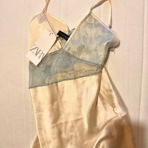 Champagnefärgad klänning från Zara - Säljer en fin champagnefärgad klänning från Zara med spetsdetaljer i ljusgrön, aldrig använd med prislapp. Klänningen har smala axelband och en omlott-design framtill. Perfekt för en sommardag eller utekväll. 