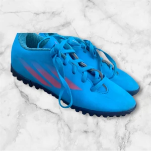 Blå fotbollsskor med rosa detaljer - Snygga blå fotbollsskor med rosa detaljer och snörning. Perfekta för spel på konstgräs med sin greppvänliga sula. Skorna har en modern design och är tillverkade i syntetmaterial för hållbarhet och komfort.