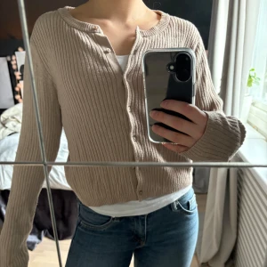 Beige ribbad kofta - Säljer en stilren beige ribbad kofta med knappar framtill. Perfekt för lager-på-lager och passar till både jeans och kjol. Koftan har långa ärmar och en klassisk rund halsringning. Ett måste i garderoben för en avslappnad look.