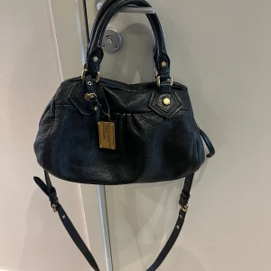 Marc Jacobs väska  - Mini modellen. Mått ca 30 cm bred och 18 cm höjd🩷 Snygg svart handväska i skinn med gulddetaljer och dragkedja. Väskan har både handtag och en justerbar axelrem. Insidan är fodrad med ett mönster av bokstäver. Perfekt för att bära dina dagliga nödvändigheter med stil.