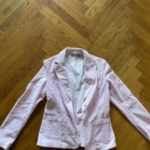 Rosa randig kavaj från Zoul - Säljer en stilren rosa kavaj från Zoul med diskreta ränder. Kavajen har en klassisk design med knappar framtill och två fickor. Perfekt för att ge en färgklick till din outfit!