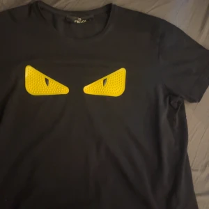 Svart t-shirt med gula detaljer  - Säljer en svart t-shirt med coola gula ögonliknande detaljer på framsidan. Perfekt för dig som vill sticka ut med en unik design. T-shirten har en klassisk passform och är kortärmad. Och ber om ursäkt för dåliga bilder.