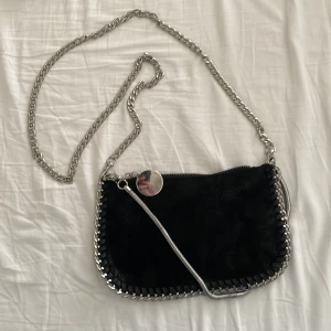 Svart axelväska med kedjedetaljer - Snygg svart axelväska med silverfärgade kedjedetaljer och dragkedja. Perfekt för att ge din outfit en edgy touch. Väskan har en lång kedjerem som gör den enkel att bära över axeln.