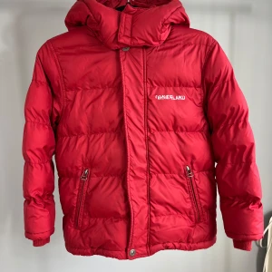 Röd dunjacka från Timberland - Snygg röd dunjacka från Timberland med dragkedja och knappar. Jackan har en stor logga på ryggen och en mindre på bröstet. Perfekt för kalla dagar med sin vadderade design och huva.