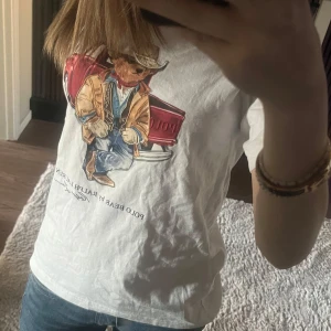 Vit t-shirt med tryck från Polo Ralph Lauren - Säljer en vit t-shirt från Polo Ralph Lauren med ett coolt tryck av en björn i cowboykläder. Perfekt för en avslappnad stil. T-shirten har en normal passform och korta ärmar.