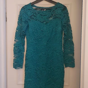 Grön spetsklänning - Snygg grön klänning i spets med långärmad design. Perfekt för en elegant look med sitt vackra blommönster. Klänningen har en tight passform som framhäver figuren.