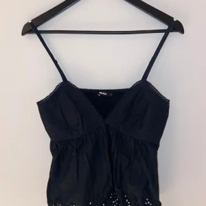 Svart linne från BikBok - Snygg svart linne från BikBok med smala axelband och v-ringning. Toppen har fina spetsdetaljer och en elastisk rygg för bättre passform. Perfekt för varma dagar!