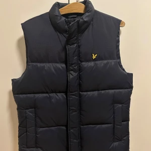 Svart dunväst från Lyle & Scott - Snygg svart dunväst från Lyle & Scott med dragkedja och broderad logga på bröstet. Perfekt för kyligare dagar och ger en stilren look. Västen har en quiltad design som håller dig varm och bekväm.