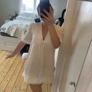 Vit klänning med spetsdetaljer - Charmig vit klänning med v-ringning och spetsdetaljer vid hals och fåll. Kortärmad design som ger en luftig och somrig känsla. Perfekt för varma dagar. Student eller sommarklänning med fina broderier