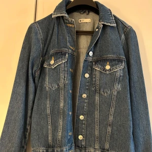 Blå jeansjacka från Gina tricot - Säljer en klassisk blå jeansjacka. Jackan har långa ärmar och knappar framtill samt två bröstfickor med knappstängning. 