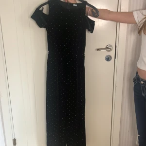 Svart långklänning med glittrigt mönster - Elegant svart långklänning med glittrigt mönster. Klänningen har en offshoulder-design som ger en stilren look. Perfekt för speciella tillfällen när du vill glänsa lite extra.